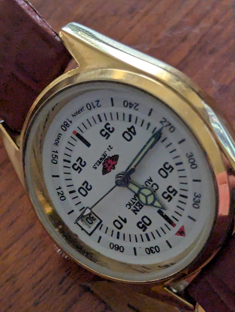 Pu&ograve; includere: Un orologio da polso dorato con quadrante bianco e cinturino in pelle marrone. Il quadrante mostra numeri, indicatori e lancette neri. L'orologio riporta la scritta "21 JEWELS AUTOMATIC JAPAN MADE".