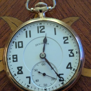 May include: A gold-tone pocket watch with a white face, black Arabic numerals, and the word ILLINOIS printed in black. The watch features a small sub-dial for seconds at the bottom, a winding crown at the top, and thin black metal hands.
