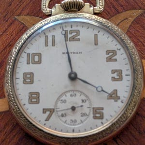 Puede incluir: Primer plano de un reloj de bolsillo Waltham dorado. La esfera blanca tiene n&uacute;meros y agujas negras. Una esfera secundaria m&aacute;s peque&ntilde;a est&aacute; en la parte inferior. El fondo es oscuro y estampado.