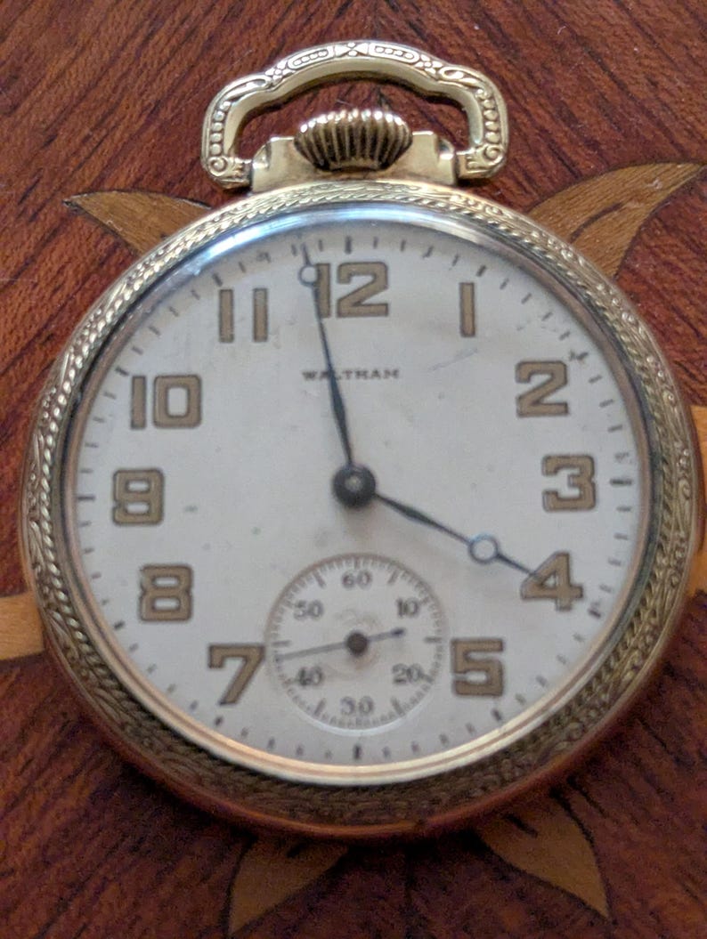 Puede incluir: Primer plano de un reloj de bolsillo Waltham dorado. La esfera blanca tiene n&uacute;meros y agujas negras. Una esfera secundaria m&aacute;s peque&ntilde;a est&aacute; en la parte inferior. El fondo es oscuro y estampado.