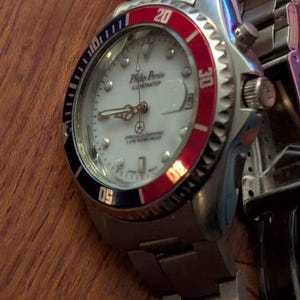 May include: A silver-tone stainless steel wristwatch with a white dial, featuring the text Philip Persio Illuminator. The watch has a rotating bezel with red and black sections, luminous hands, and a metal link bracelet with a fold-over clasp.