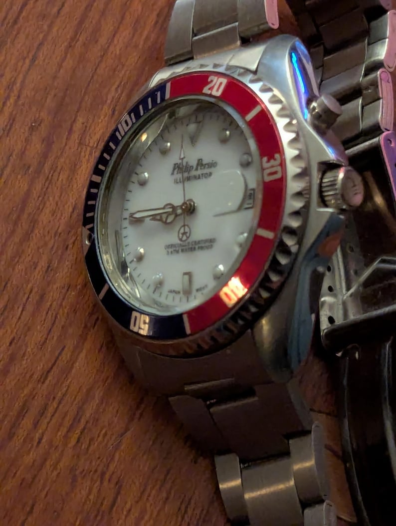 May include: A silver-tone stainless steel wristwatch with a white dial, featuring the text Philip Persio Illuminator. The watch has a rotating bezel with red and black sections, luminous hands, and a metal link bracelet with a fold-over clasp.