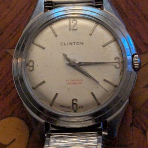 May include: A vintage Clinton wristwatch with a round silver-tone case and a white dial. The dial features silver-tone hour markers, numerals at 3, 6, 9, and 12, and the text Clinton, 17 Jewels, Incabloc, and Swiss Ed. It has a textured metal band.