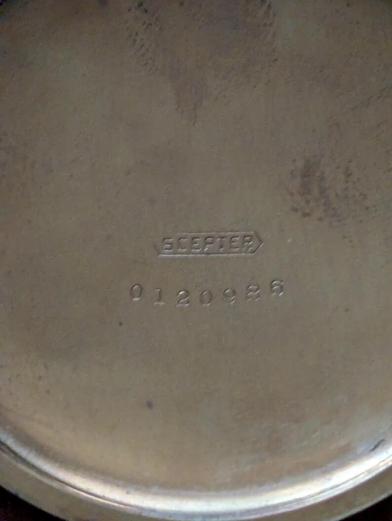 Puede incluir: Primer plano de un objeto met&aacute;lico, posiblemente de lat&oacute;n, con la palabra "Scepter" y el n&uacute;mero "0120986" grabados en la superficie. El objeto tiene forma circular con un acabado ligeramente texturizado.