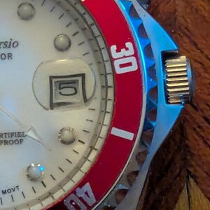 May include: A wristwatch with a white dial, silver-tone hour markers, and a date window displaying the number 5. The watch features a rotating bezel with a red insert, white numerals 30 and 40, and a metallic crown on the right side of the case.