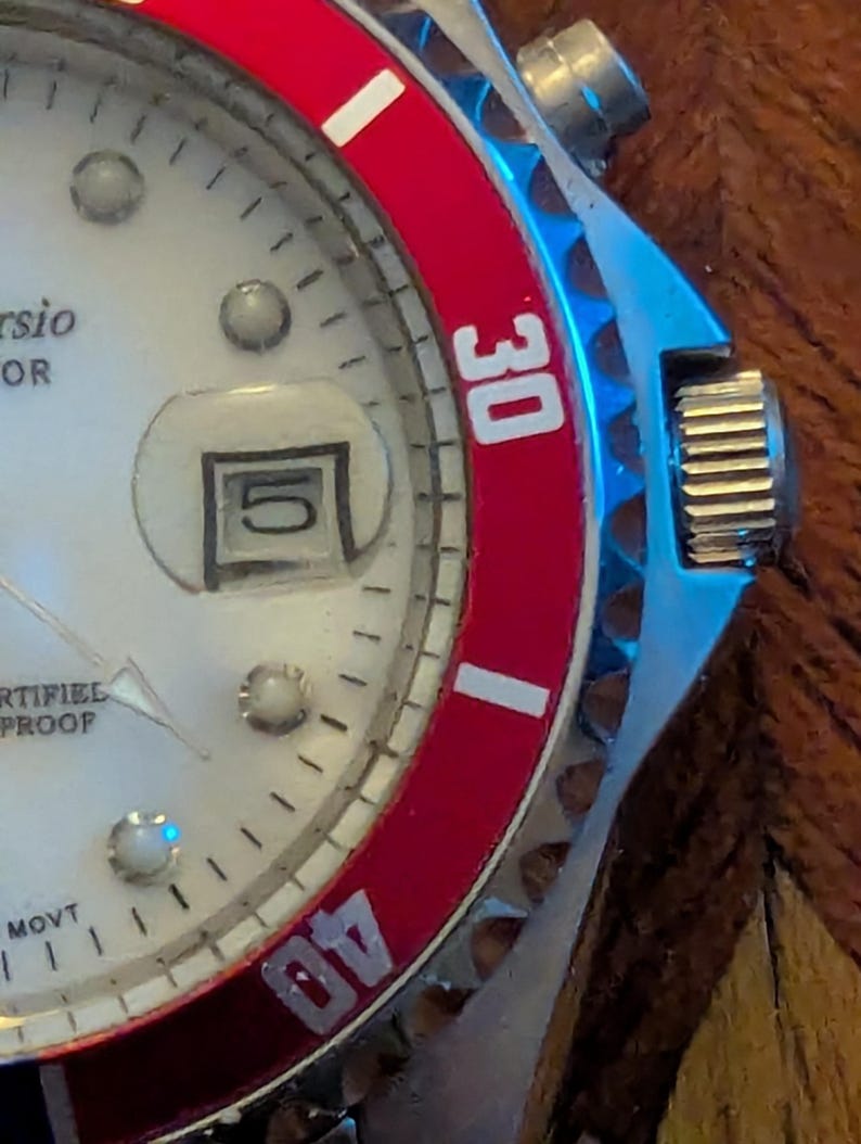 May include: A wristwatch with a white dial, silver-tone hour markers, and a date window displaying the number 5. The watch features a rotating bezel with a red insert, white numerals 30 and 40, and a metallic crown on the right side of the case.