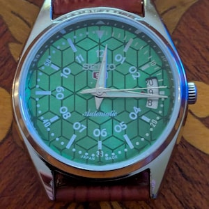 May include: A Seiko 5 automatic wristwatch with a silver-tone stainless steel case and a brown leather strap. The green dial features a geometric hexagonal pattern, white Arabic numerals, silver hands, and a date window at the three o'clock position.