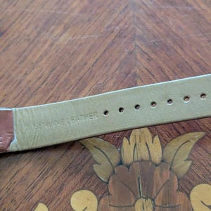 Pu&ograve; includere: Primo piano di un cinturino per orologio in pelle marrone con le parole "GENUINE LEATHER" stampate sul lato inferiore. Il cinturino ha diversi fori per la regolazione. Lo sfondo &egrave; una superficie di legno con un disegno floreale.