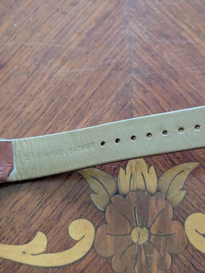 Pu&ograve; includere: Primo piano di un cinturino per orologio in pelle marrone con le parole "GENUINE LEATHER" stampate sul lato inferiore. Il cinturino ha diversi fori per la regolazione. Lo sfondo &egrave; una superficie di legno con un disegno floreale.