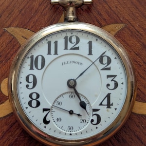 May include: A gold-tone pocket watch with a white dial, black Arabic numerals, and the word ILLINOIS printed in the center. The watch features a small sub-dial for seconds at the bottom and a winding crown at the top with a circular bow.