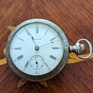 May include: A silver pocket watch with Roman numeral markings on the face. The watch face is white with black markings and the word "ALTHAM" printed in blue. The watch has a decorative engraved border.