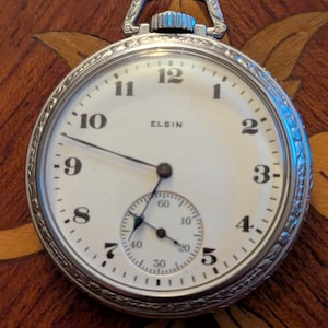 May include: A silver-toned Elgin pocket watch with a white face and black numerals. The watch has a subdial and a decorative bezel. The word "ELGIN" is printed on the watch face.