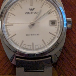 May include: A vintage Waltham self-winding wristwatch. The watch features a silver-colored case and a white face with black hour markers and hands. The watch has a date display at the 3 o'clock position and a black and silver band. The watch is a classic timepiece.