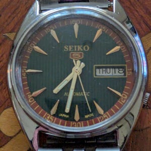 May include: A silver-toned Seiko 5 automatic wristwatch with a dark green face and a red and gold outer ring. The watch face displays the day and date, with gold hands and markers. The watch band is silver-toned.