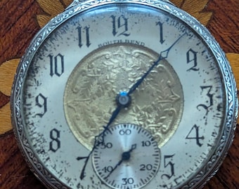 Puede incluir: Un reloj de bolsillo plateado con una esfera blanca y agujas negras. El reloj tiene un pequeño subdial blanco redondo con agujas negras y marcas negras. La esfera del reloj tiene marcadores de hora romanos negros y las palabras "SOUTH BEND" impresas en él.