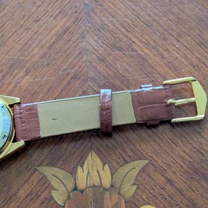 Pu&ograve; includere: Primo piano di un orologio da polso dorato con cinturino in pelle marrone. Il cinturino presenta una texture effetto coccodrillo e una fibbia dorata. L'orologio &egrave; appoggiato su una superficie in legno con intarsi floreali.