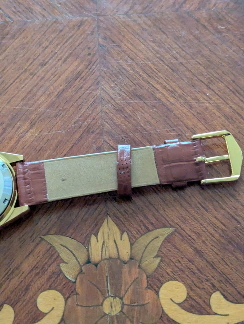 Pu&ograve; includere: Primo piano di un orologio da polso dorato con cinturino in pelle marrone. Il cinturino presenta una texture effetto coccodrillo e una fibbia dorata. L'orologio &egrave; appoggiato su una superficie in legno con intarsi floreali.