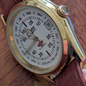 Pu&ograve; includere: Un orologio da polso automatico Citizen color oro con quadrante bianco, numeri e indicatori neri. L'orologio ha un cinturino in pelle marrone e un datario alle 3. Il quadrante recita "21 Jewels".