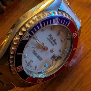 May include: A silver-tone analog wristwatch with a white dial, featuring the text Philip Persio Illuminator. The bezel is divided into red and blue sections with white numerals. It includes a date window, luminous hands, and a metal link band.