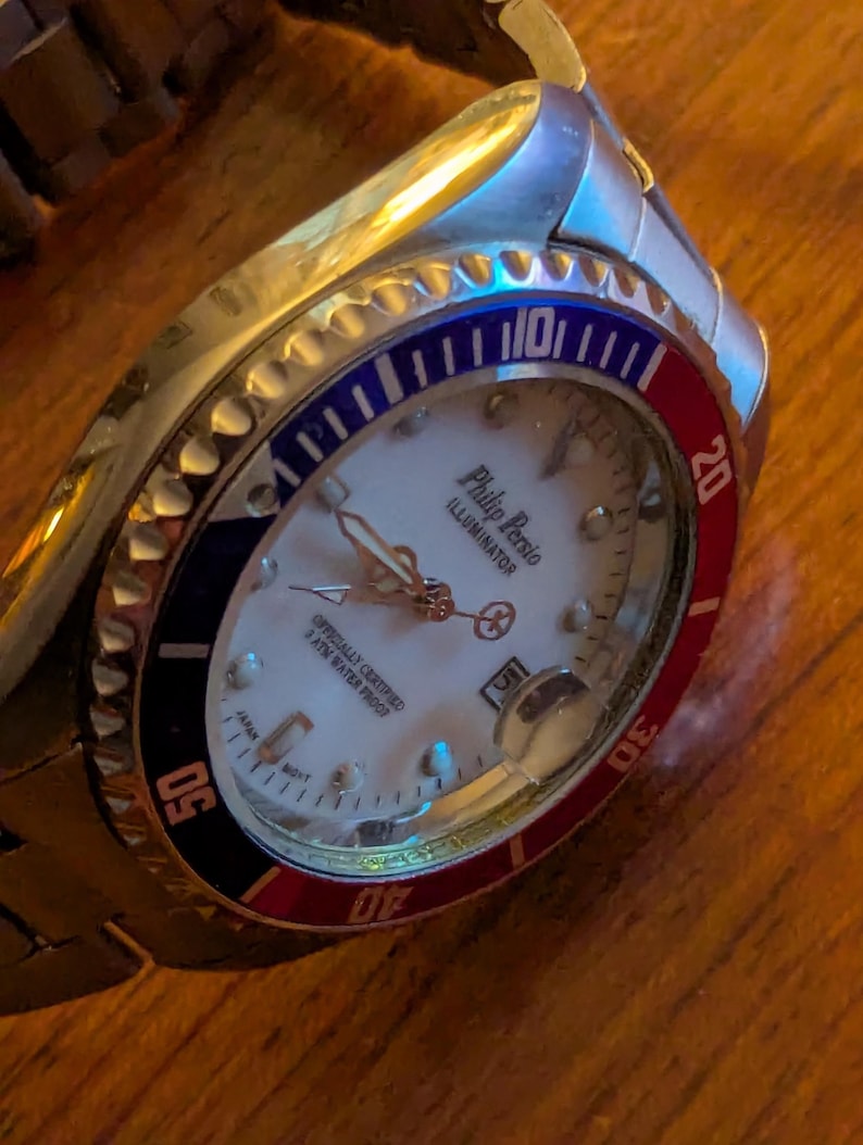 May include: A silver-tone analog wristwatch with a white dial, featuring the text Philip Persio Illuminator. The bezel is divided into red and blue sections with white numerals. It includes a date window, luminous hands, and a metal link band.