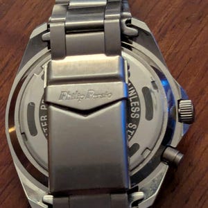 May include: A silver-toned stainless steel wristwatch with a metal link bracelet. The back of the watch case and the clasp are visible, featuring the engraved text Philip Persio and Stainless Steel. The watch has a circular case and a side crown.