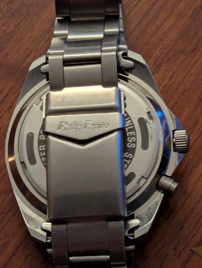 May include: A silver-toned stainless steel wristwatch with a metal link bracelet. The back of the watch case and the clasp are visible, featuring the engraved text Philip Persio and Stainless Steel. The watch has a circular case and a side crown.