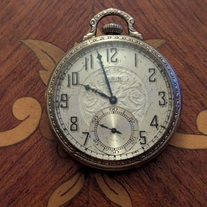 May include: An antique Elgin pocket watch with a gold-tone case and a white face. The watch features black numerals, a subdial, and ornate detailing on the face. The watch is set against a wooden surface with decorative inlays.