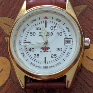 Pu&ograve; includere: Orologio automatico Citizen color oro con quadrante bianco, numeri neri e cinturino in pelle marrone. Il quadrante mostra la data e le parole "Citizen Automatic" sono visibili. L'orologio ha un marchio di 21 rubini.