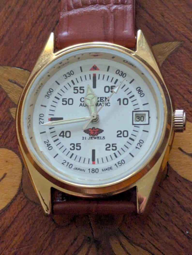 Pu&ograve; includere: Orologio automatico Citizen color oro con quadrante bianco, numeri neri e cinturino in pelle marrone. Il quadrante mostra la data e le parole "Citizen Automatic" sono visibili. L'orologio ha un marchio di 21 rubini.