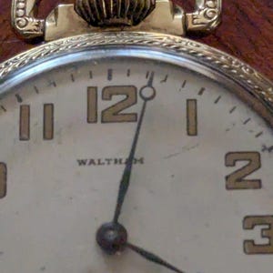 Puede incluir: Primer plano de un reloj de bolsillo Waltham vintage en tono dorado. La esfera es blanquecina con manecillas negras y n&uacute;meros dorados. El reloj tiene una parte superior decorativa y un peque&ntilde;o dial de segundos.