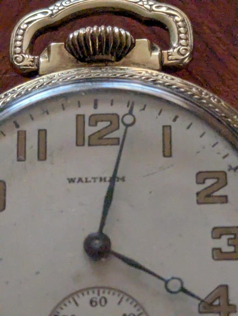 Puede incluir: Primer plano de un reloj de bolsillo Waltham vintage en tono dorado. La esfera es blanquecina con manecillas negras y n&uacute;meros dorados. El reloj tiene una parte superior decorativa y un peque&ntilde;o dial de segundos.