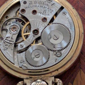 Puede incluir: Primer plano del mecanismo de un reloj de bolsillo dorado vintage. La esfera del reloj revela intrincados engranajes, resortes y el texto "A.W.W.Co. Waltham, Mass. U.S.A. 17 Jewels". El reloj tiene una caja dorada y un lazo decorativo.