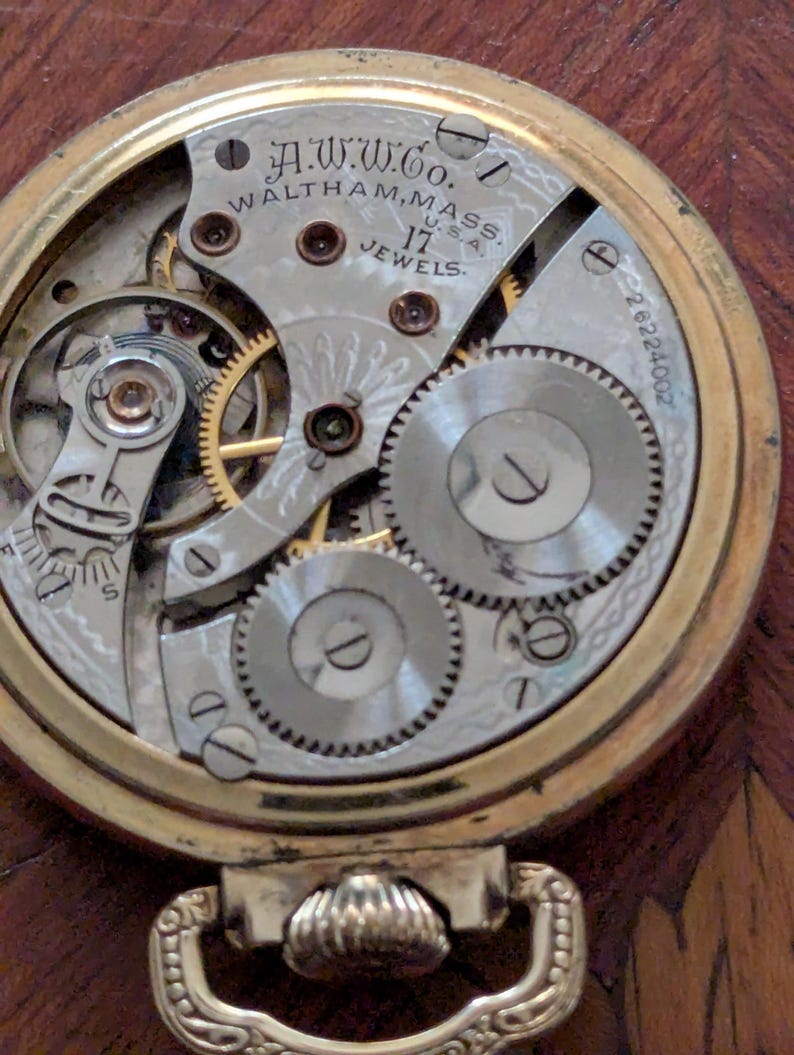 Puede incluir: Primer plano del mecanismo de un reloj de bolsillo dorado vintage. La esfera del reloj revela intrincados engranajes, resortes y el texto "A.W.W.Co. Waltham, Mass. U.S.A. 17 Jewels". El reloj tiene una caja dorada y un lazo decorativo.