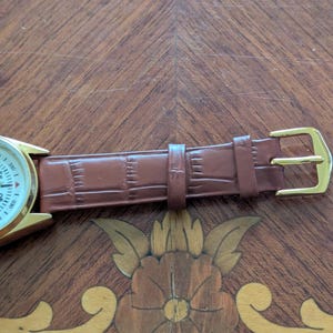 Pu&ograve; includere: Un orologio da polso dorato con quadrante bianco e cinturino in pelle marrone. Il cinturino presenta un motivo strutturato e una fibbia dorata. L'orologio &egrave; esposto su una superficie di legno con intarsi floreali.