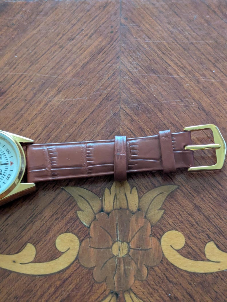 Pu&ograve; includere: Un orologio da polso dorato con quadrante bianco e cinturino in pelle marrone. Il cinturino presenta un motivo strutturato e una fibbia dorata. L'orologio &egrave; esposto su una superficie di legno con intarsi floreali.