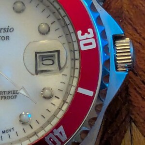 May include: A wristwatch with a white dial, silver-tone hands, and a date window displaying the number 5. The watch features a red rotating bezel with white numerals 30 and 40, a silver-tone metal case, and a textured crown on the right side.