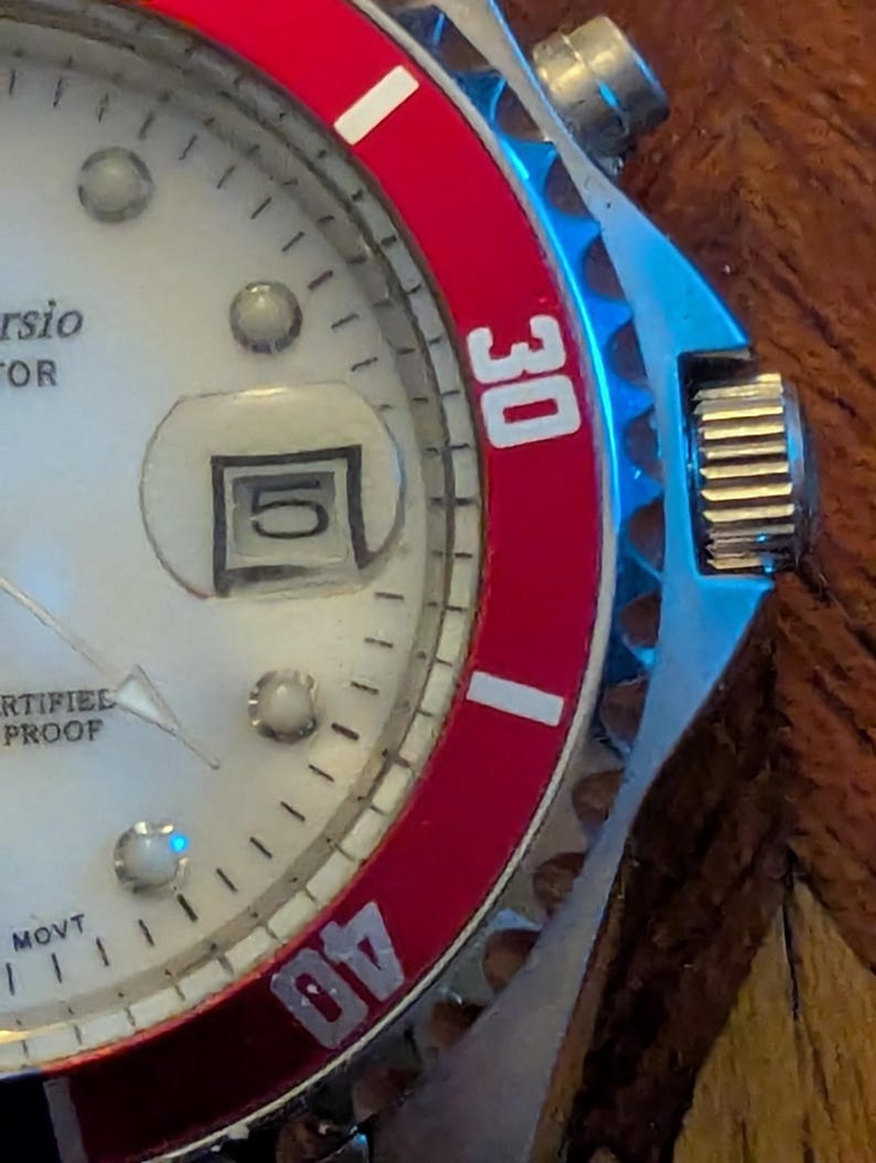 May include: A wristwatch with a white dial, silver-tone hands, and a date window displaying the number 5. The watch features a red rotating bezel with white numerals 30 and 40, a silver-tone metal case, and a textured crown on the right side.