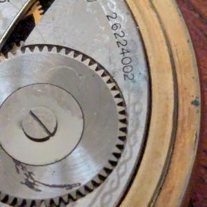 Puede incluir: Primer plano del mecanismo de un reloj antiguo. La imagen muestra los intrincados detalles de los componentes internos del reloj, incluyendo engranajes, tornillos y un n&uacute;mero de serie. El reloj tiene un marco dorado y una esfera plateada.