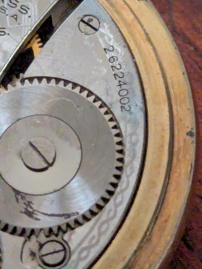 Puede incluir: Primer plano del mecanismo de un reloj antiguo. La imagen muestra los intrincados detalles de los componentes internos del reloj, incluyendo engranajes, tornillos y un n&uacute;mero de serie. El reloj tiene un marco dorado y una esfera plateada.