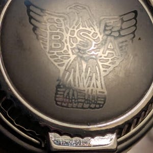 May include: A circular silver-toned metal object featuring a dark gray center inlay. The inlay displays an engraved eagle emblem with the letters BSA positioned across the bird's chest. The outer rim has a textured, ridged edge with a small metal tab.