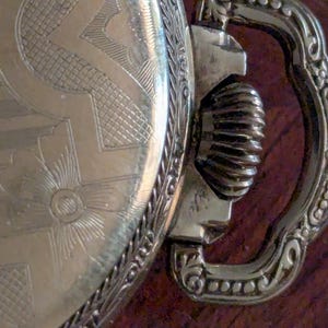 Puede incluir: Primer plano de un reloj de bolsillo vintage. El reloj presenta un dise&ntilde;o grabado y ornamentado en la esfera y un asa decorativa. El metal tiene un tono dorado y el reloj muestra signos de antig&uuml;edad.
