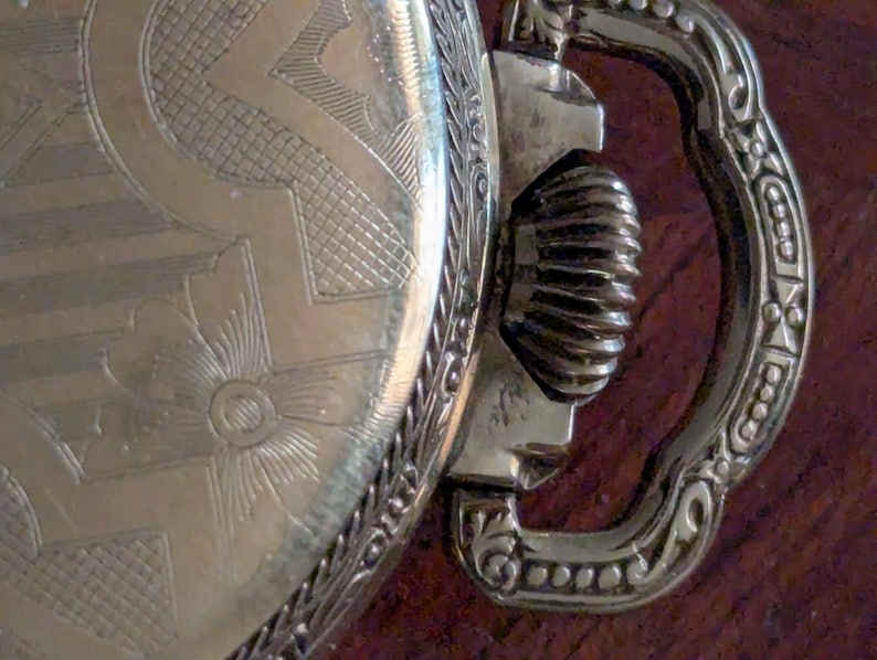 Puede incluir: Primer plano de un reloj de bolsillo vintage. El reloj presenta un dise&ntilde;o grabado y ornamentado en la esfera y un asa decorativa. El metal tiene un tono dorado y el reloj muestra signos de antig&uuml;edad.