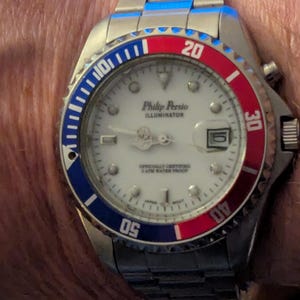May include: A Philip Persio Illuminator wristwatch with a white dial, silver-tone hands, and a date window. The bezel features a blue and red color scheme with white numerals. The watch has a stainless steel link bracelet and a round case.