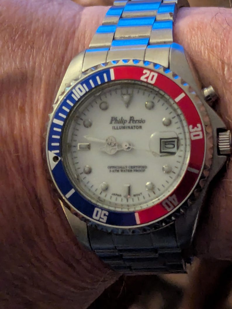 May include: A Philip Persio Illuminator wristwatch with a white dial, silver-tone hands, and a date window. The bezel features a blue and red color scheme with white numerals. The watch has a stainless steel link bracelet and a round case.