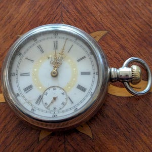 May include: A silver pocket watch with a white face, black Roman numerals, and a smaller seconds dial. The watch has gold-colored hands and a winding stem. The watch is set on a wooden surface.