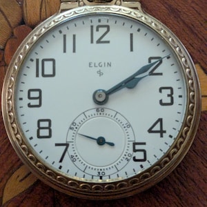 May include: A gold-tone pocket watch with a white dial, black Arabic numerals, and dark blue hands. The watch face features the text ELGIN and a small sub-dial for seconds at the bottom. The outer case has a decorative scalloped edge pattern.