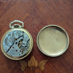 Puede incluir: Reloj de bolsillo abierto con engranajes expuestos y una caja dorada. El mecanismo del reloj es visible, con intrincados engranajes y las palabras "Waltham, Mass." y "17 Jewels". El reloj est&aacute; abierto, con la tapa trasera al lado.