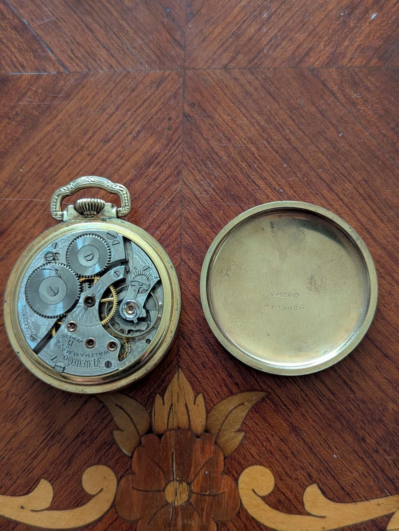 Puede incluir: Reloj de bolsillo abierto con engranajes expuestos y una caja dorada. El mecanismo del reloj es visible, con intrincados engranajes y las palabras "Waltham, Mass." y "17 Jewels". El reloj est&aacute; abierto, con la tapa trasera al lado.