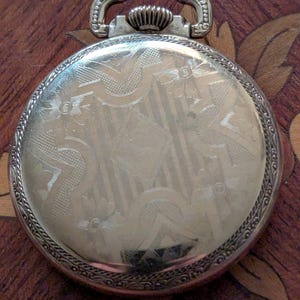 Puede incluir: Un reloj de bolsillo plateado con un dise&ntilde;o ornamentado en la cubierta trasera. La cubierta presenta un patr&oacute;n grabado detallado con elementos florales y geom&eacute;tricos. El reloj tiene un asa decorativa y una corona texturizada. El reloj est&aacute; sobre una superficie de madera.