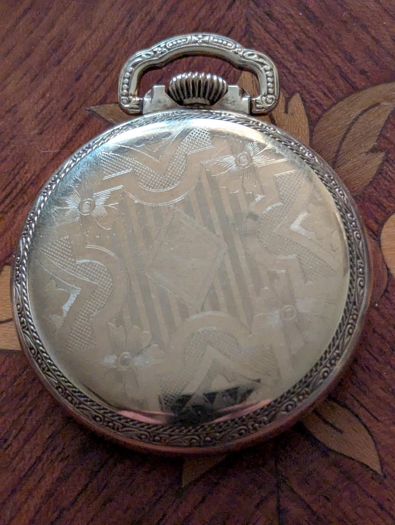 Puede incluir: Un reloj de bolsillo plateado con un dise&ntilde;o ornamentado en la cubierta trasera. La cubierta presenta un patr&oacute;n grabado detallado con elementos florales y geom&eacute;tricos. El reloj tiene un asa decorativa y una corona texturizada. El reloj est&aacute; sobre una superficie de madera.